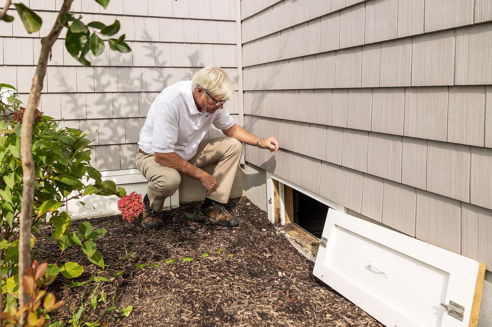 A homeowner crouched at the open crawl-space entrance of a house during an inspection