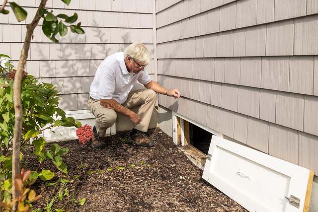 A homeowner crouched at the open crawl-space entrance of a house during an inspection
