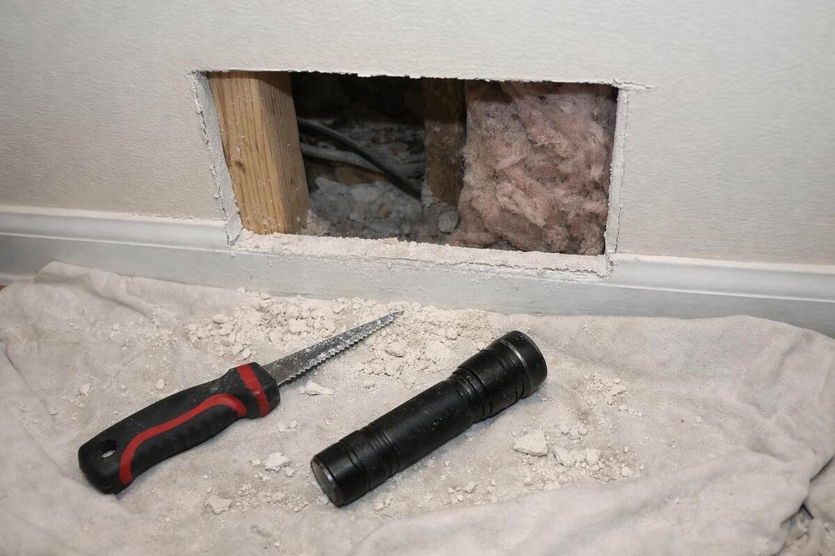 A rectangular access opening cut into hallway drywall with a jab saw and flashlight on the floor below